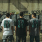 Três homens de costas vestindo t-shirts astredik com ilustração de lata de spray azul em fundo de parede com graffiti.