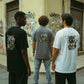 Três homens vestem t-shirts astredik com estampa de caveira estilo graffiti em fundo urbano.